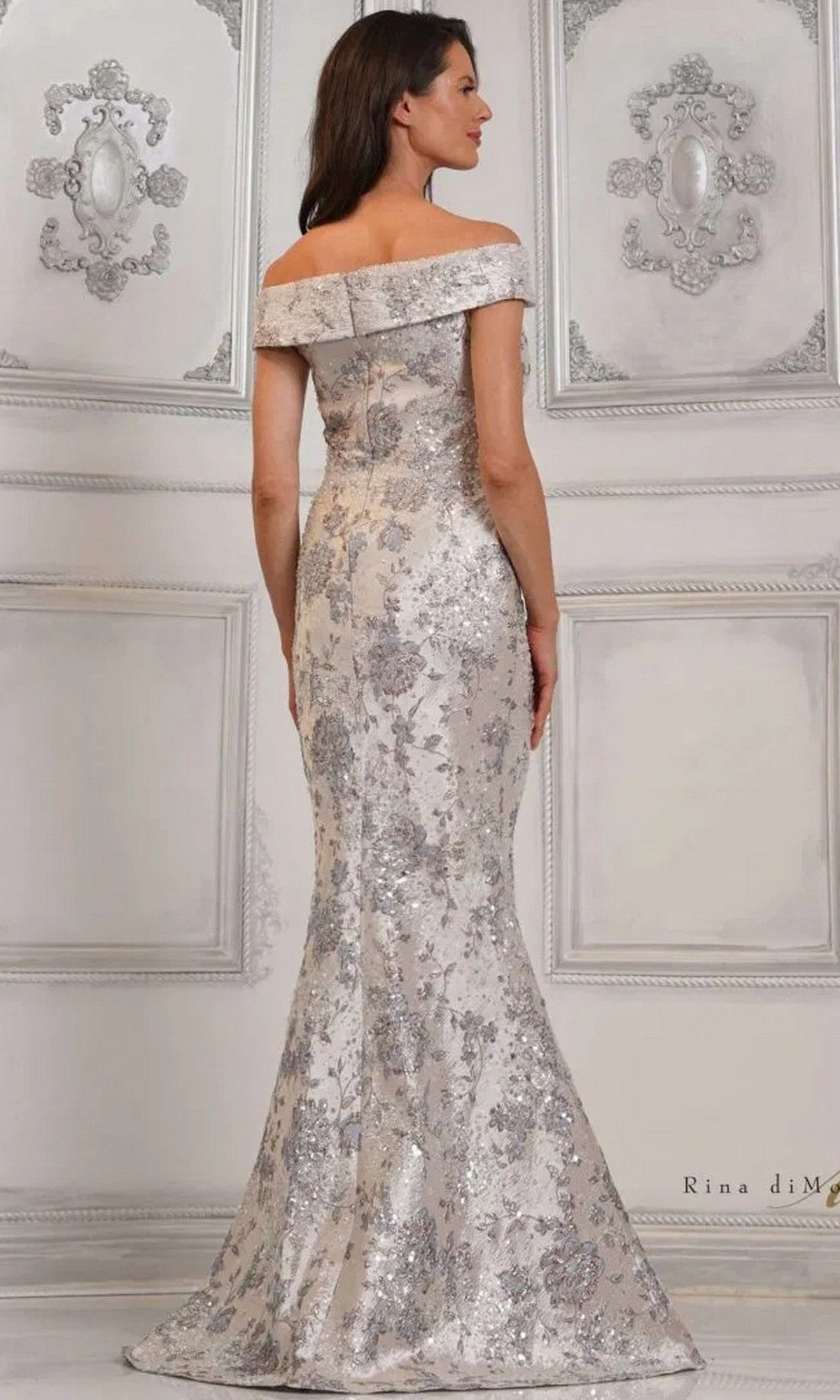 A woman faces away, wearing the Rina Di Montella RD3106—an off-the-shoulder, floor-length mermaid formal dress in silver and white floral beaded jacquard brocade—set in an ornate, light-colored room.